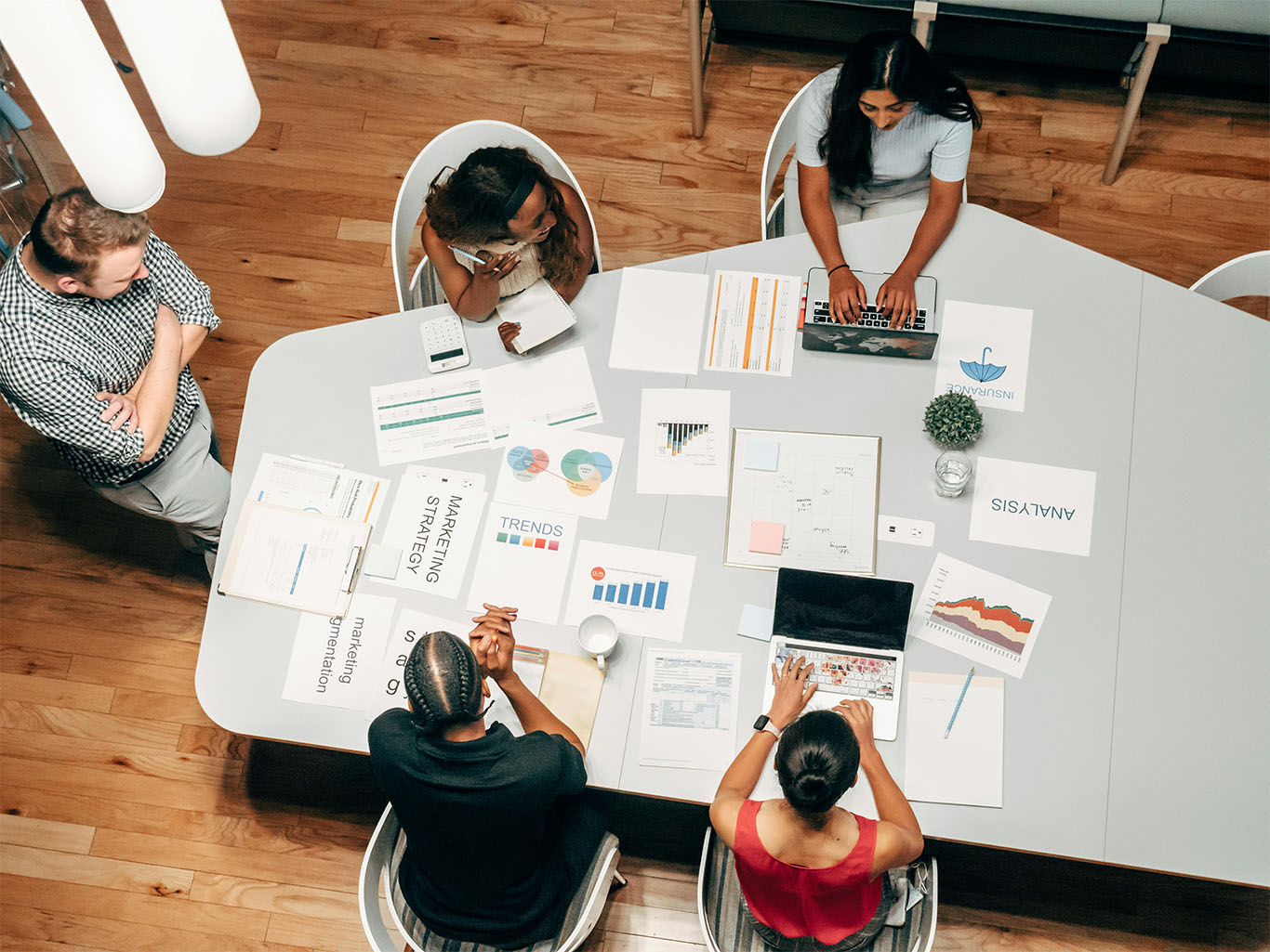 A diverse group of professionals sitting around a conference table reviewing marketing strategy documents, trend graphs, and analytics reports during a team meeting.