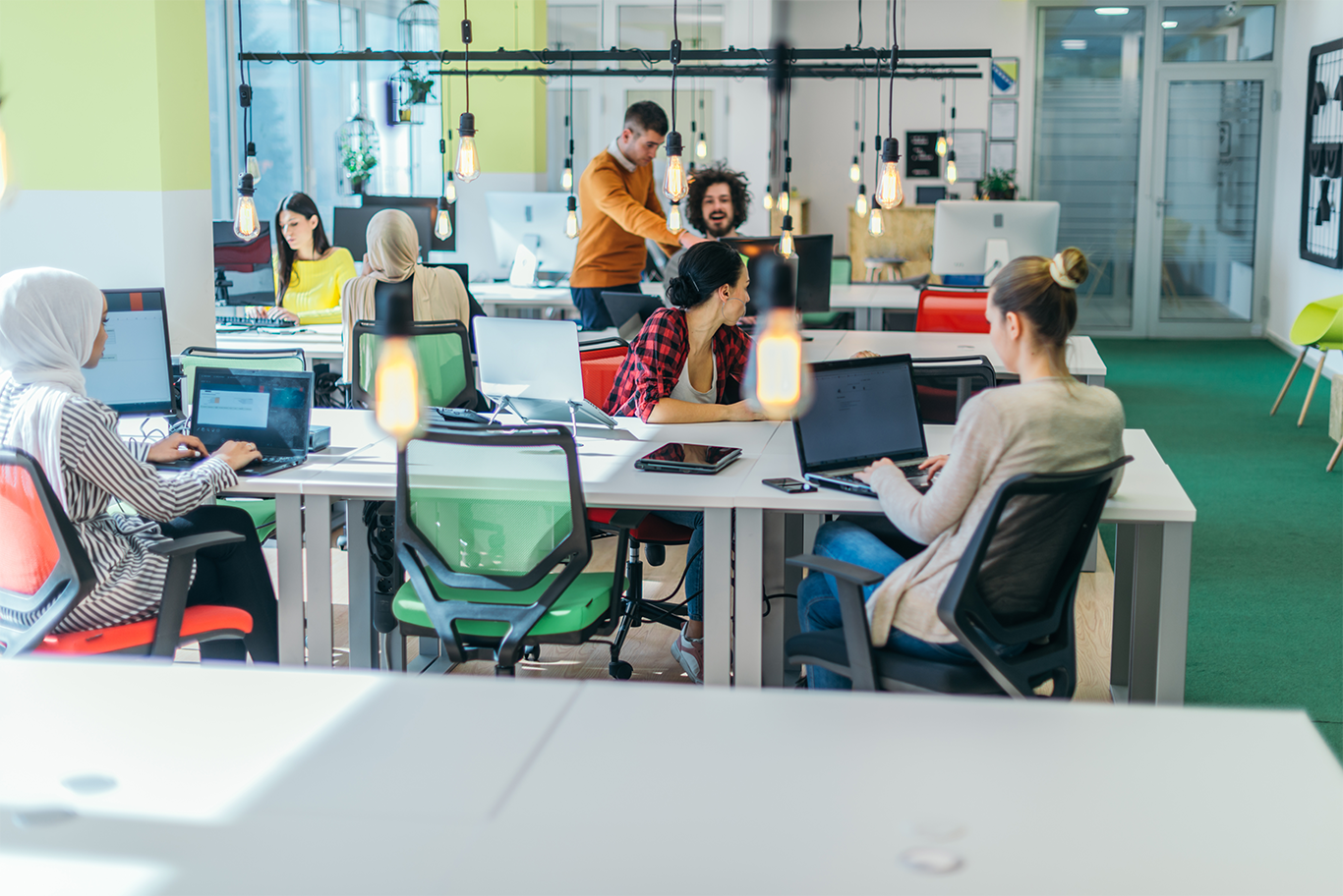 A modern open-plan office where a team of marketing professionals work collaboratively at desks with laptops and desktop computers in a bright, contemporary workspace.
